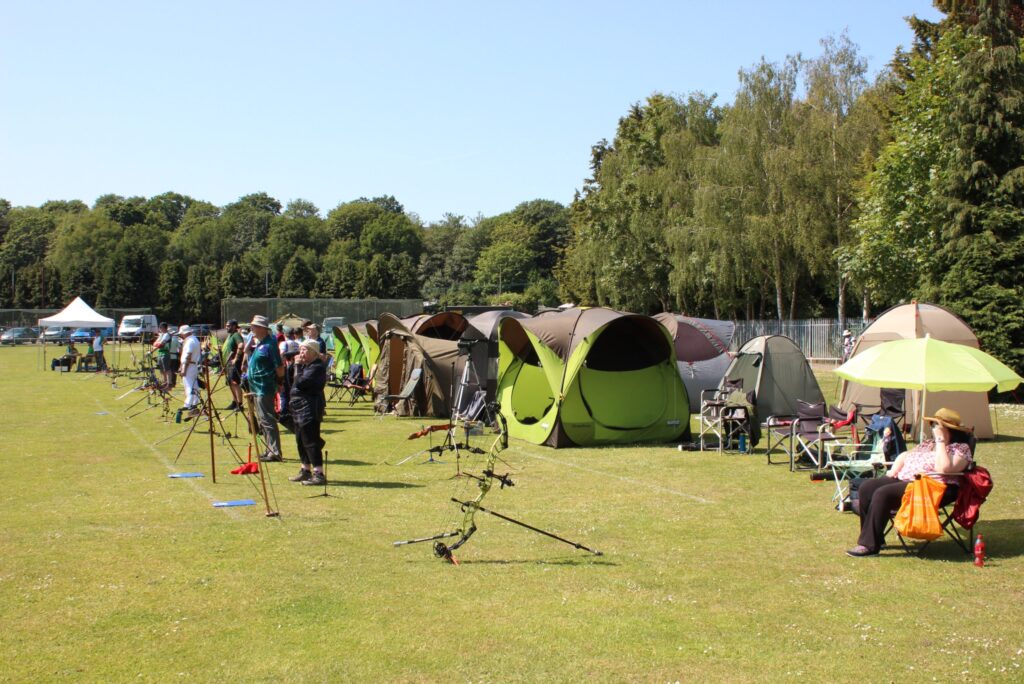 A photograph of various archers waiting by the shooting line in front of tents and chairs.